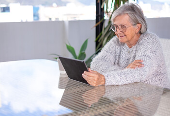 Senior attractive woman using digital tablet sitting outdoor on a large table, smiling grey-haired woman looking at web pages enjoying tech and social
