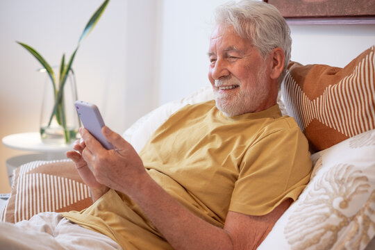 Smiling relaxed senior man lying in bed using a smartphone communicating in remote chat with family or browsing internet