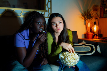 Two young women, an interracial lesbian couple, are sitting on a sofa at home, eating popcorn and...