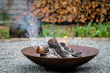 A close-up shot of burning firewood in a shallow, rusted steel fire pit placed on a gravel surface. Light smoke rises into the air while small flames dance around the blackened logs. 
