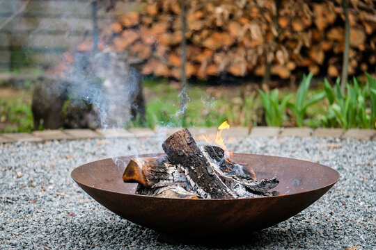 A close-up shot of burning firewood in a shallow, rusted steel fire pit placed on a gravel surface. Light smoke rises into the air while small flames dance around the blackened logs. 