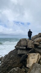 Solitary figure in a dark jacket gazing at the ocean from rocks, symbolizing introspection and...