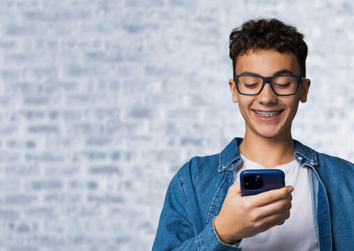 Smiling black curly hair funny young man, guy, boy wear braces hold hand typing text, looking reading gazing at cell phone mobile smartphone cellphone, stand against white bricks loft wall background