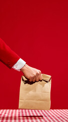 Hand in red suit holding kraft paper bag over red checkered table against vivid red background....