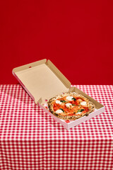 Open pizza box with delicious pizza on red checkered tablecloth against bold red background. Concept of food, pop art photography, hunger, visual satisfaction, food delivery services, restaurant menu