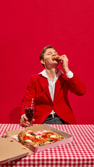 Man in red suit with eyes closed enjoying pizza slice with soda on checkered table against bright red background. Concept of food, emotions, pop art, indulgence, pleasure, color, and lifestyle.