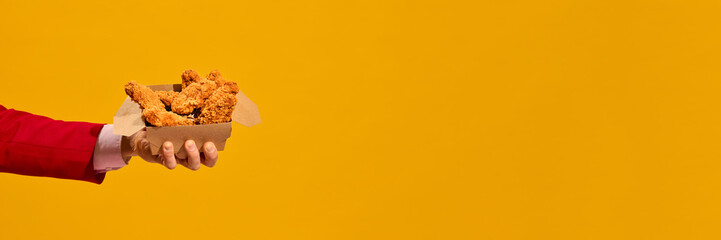 Hand in red sleeve holding box of crispy fried chicken nuggets against vibrant yellow background....