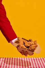 Hand in red sleeve holding box of crispy fried chicken nuggets against yellow background over checkered tablecloth. Concept of fast food aesthetics, pop art, restaurant menu. Vertical image