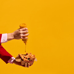 Hand squeezing mustard sauce bottle over crispy fried chicken nuggets against bold yellow background. Spicy taste of fast food. Concept of taste explosion, contrast, food art photography