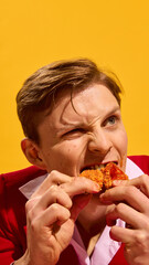 Close-up portrait of man emotionally biting crispy chicken wing with sauce on lips and intense expression against yellow background. Concept of fast food aesthetics, pop art photography, emotions