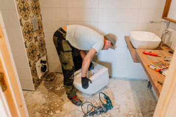 A construction worker installs a toilet in a bathroom under renovation, with tools and debris...