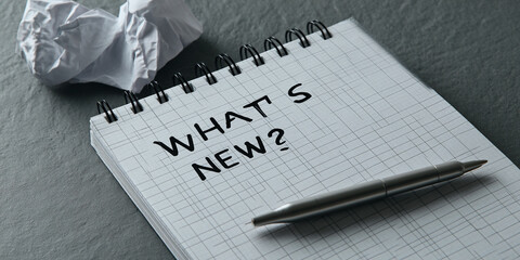 Spiral notepad with the words "What's New?" written on it, pen beside it, crumpled paper in background, showcasing a concept of brainstorming, news, updates or new ideas