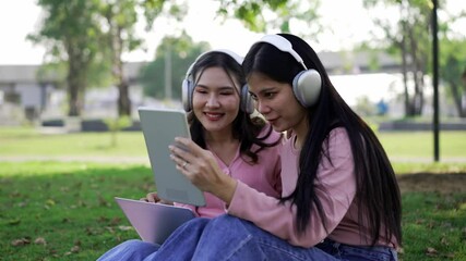 Locked medium shot, Young asian Lesbian Couple sharing loving moment, Enjoying with music through headphone, use laptop and tablet, talking and smile with happy during relaxation at public park - Powered by Adobe
