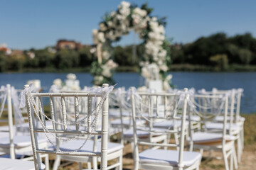 A wedding ceremony with white chairs, a path and an arch of white flowers on the lake shore.