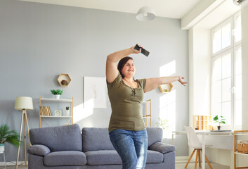 Young fat happy smiling girl wearing casual clothes dancing with smartphone in hand in the living room at home. Plus size overweight woman having fun with a satisfied face expression.