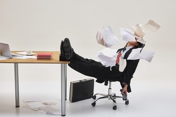 Man sitting in chair with legs on desk, surrounded by flying papers in chaotic office mess,...
