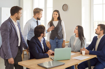 Diverse group of business professionals engaged in discussion around desk in modern office. Attentive company employees listening to man colleague or leader suggesting ideas during work meeting.
