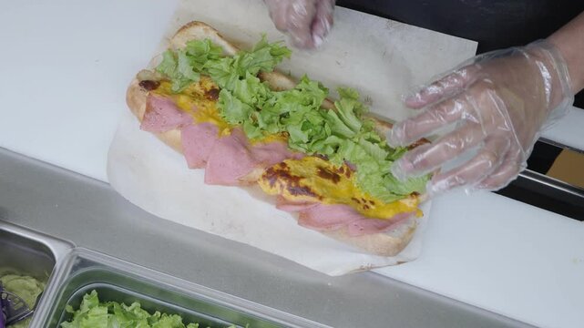 Gloved hands add lettuce to ham and omelette sub sandwich at prep counter.