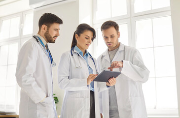 Three professional doctors gathered in medical office looking at tablet screen and discussing...