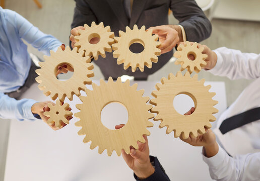 In office environment, several business people hold various wooden gears together over meeting table. Colleagues demonstrate importance of teamwork and strategic planning in business operations.