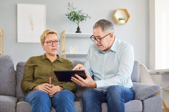 Senior couple sitting on sofa in living room at home and using tablet together browsing apps. Elderly man and woman surfing social media on digital gadget. People and technology concept.
