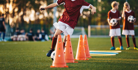 Young soccer players in maroon jerseys practice dribbling through cones on a green field during a training session at sunset