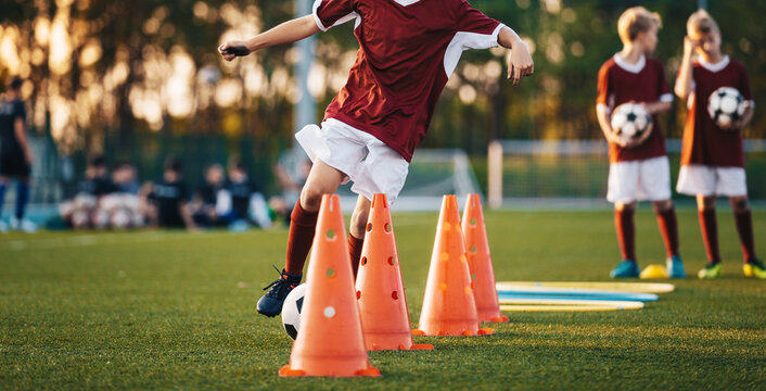 Young soccer players in maroon jerseys practice dribbling through cones on a green field during a training session at sunset