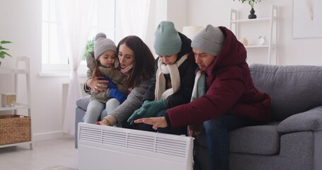 Upset and frozen parents and two kids, dressed in outerwear, sit at home near a heater trying to stay warm during freezing winter weather, showcasing family resilience against the cold.