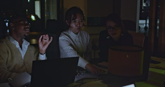 Three diverse people work late in a modern office, collaborating on a project using laptops and paperwork under mood lighting.