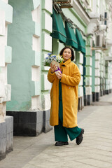 Young beautiful asian lady holding a branch of yellow flowers. She smiles widely, wearing bright clothes on the city street