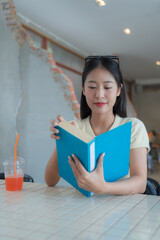 Young asian student is enjoying reading a book while sitting at a table with a refreshing drink in...