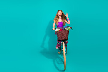 Joyful young woman riding a decorative bicycle with a flower basket against a vibrant teal background, exuding cheerful energy