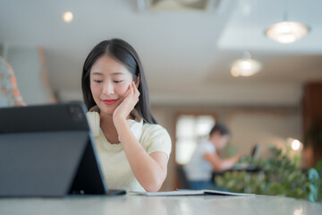 Young Asian businesswoman using tablet and working in modern office, headphones around her neck, a notebook on the desk and a colleague working in the background