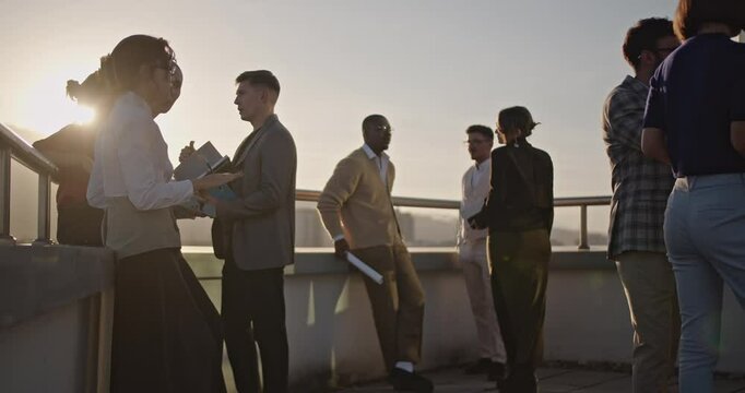 Business professionals gather on a high-rise rooftop at sunset, discussing work, collaborating, and enjoying city views.