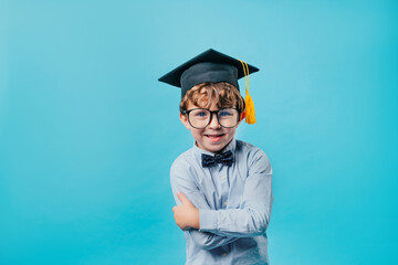 Emotional preschool boy wearing graduation cap, glasses and bow tie over blue background