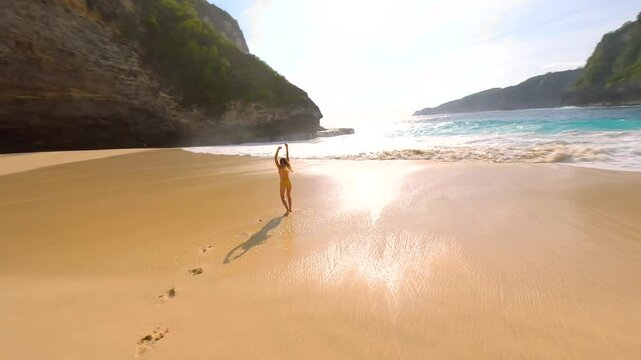 FPV drone shot of joyful woman in yellow swimsuit on tropical beach with turquoise sea, waves, and cliffs, leaving footprints in the sand, Bali, Indonesia. Concept of freedom, travel, summer vacation.
