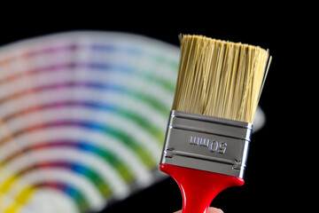 A paintbrush held in front of a color swatch fan deck displaying a full color palette. Commonly used in painting, design, and interior decoration for selecting and matching colors.