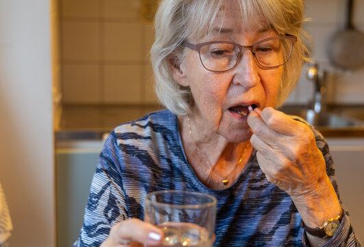 An elderly, gray-haired woman takes her medication, holding a glass of water in one hand and a pill in the other.