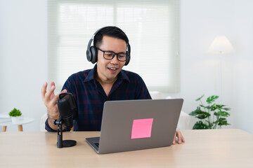 Young man in headphones engaging in virtual meeting while using laptop and microphone in bright indoor space with plants and natural lighting