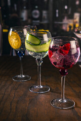 Three stylish cocktails served in large glasses with orange, cucumber and red berries, standing on a wooden surface in a moody bar ambiance, great for concepts of drinks, nightlife or bar aesthetics.