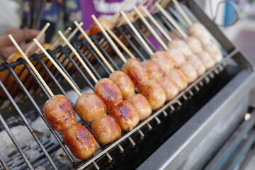 Grilled fermented sausages on skewers being cooked over charcoal at a Thai street food stall. The glossy, golden-brown sausages are a popular snack known as 