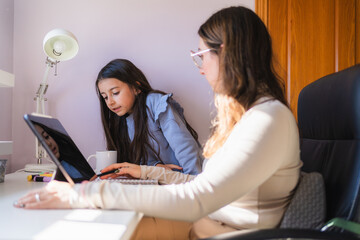 Woman assisting her young daughter with online education using a tablet, working together at a home office desk