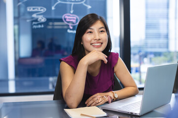 Asian woman working on laptop at glass desk in office, showing marker diagrams and city view