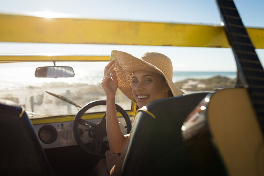 woman sitting in yellow dune buggy at sandy beach, adjusting straw hat and reaching toward guitar - Powered by Adobe