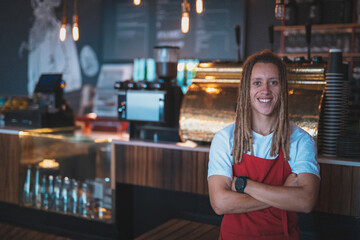 Young adult male barista standing smiling behind counter in cafe with steaming espresso machine