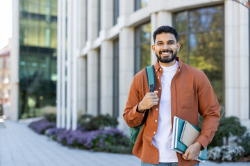 A smiling student poses outside a modern building, holding books and a backpack, ready for classes...