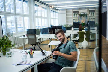 Man sitting and reviewing DSLR camera images at modern office desk, with tablet, prints, copy space