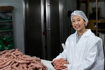 Asian woman handling tray of raw sausages on table in meat plant, wearing hairnet, copy space