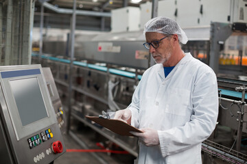 Senior QC inspector inspecting control panel at beverage factory holding clipboard near bottle belt