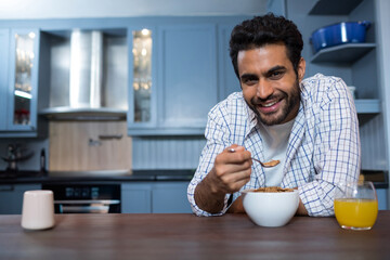 Young Indian man sitting at kitchen island eating cereal, with spoon and orange juice, copy space